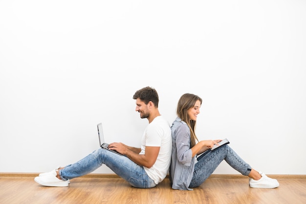 Photo the smile couple sit with a laptop and a tablet near the empty wall