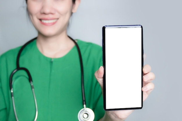 Il sorriso e l'infermiera felice indossano un abito verde con lo stetoscopio sulla spalla e mostrano lo smartphone con schermo bianco che introduce l'assistenza sanitaria tramite un'applicazione digitale