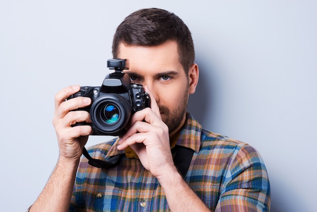 Улыбка! Портрет уверенного в себе молодого человека в рубашке, держащего камеру перед его лицом, стоя на сером фоне