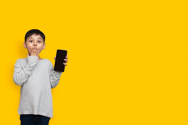 Photo smile and shocked little boy kid in green grey shirt blank screen of mobile phone on yellow background copy space