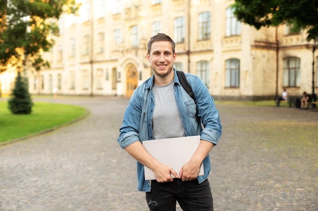 Smile student man is standing with backpack and laptop behind park near campus.