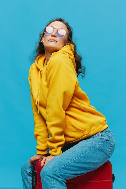 Smile woman sitting on a suitcase in a yellow hoodie blue jeans and glasses on a blue background packing for a trip
