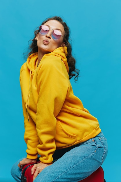 Smile woman sitting on a suitcase in a yellow hoodie blue jeans and glasses on a blue background packing for a trip