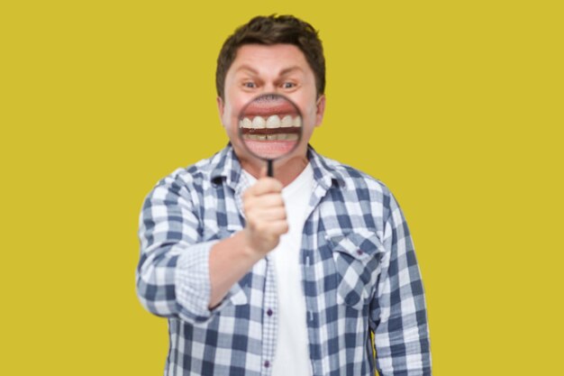 Smile zoom of tand zorg concept. Portret van een man van middelbare leeftijd in een casual geruit hemd dat staat, vergrootglas op zijn tanden houdt en naar de camera kijkt. studio opname, geïsoleerd op gele achtergrond.