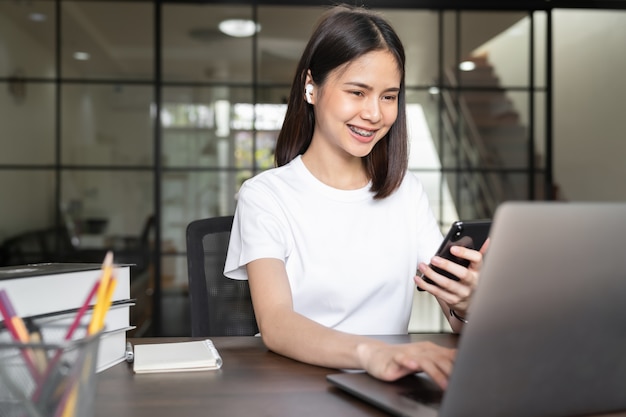 Smiley Asian woman hand holding smartphone and press the button on the keyboard laptop in house.