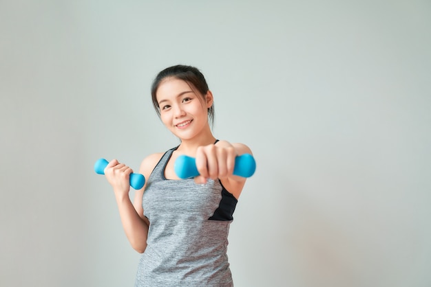 Smiley Asian woman wearing sportswear pumping up muscles with blue dumbbell . Healthy lifestyle concept.