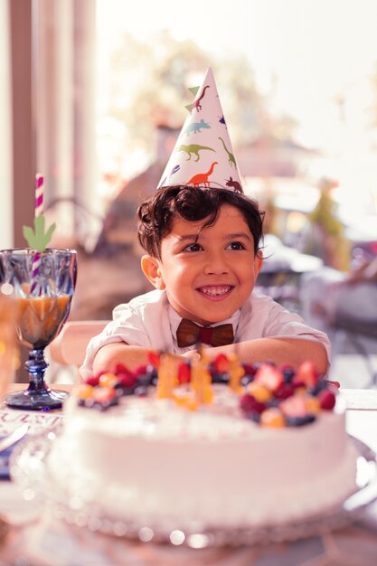 Smiley boy wearing party hat while sitting next to his birthday cake