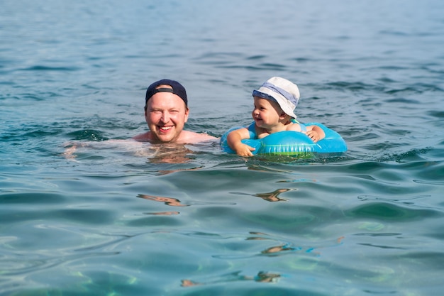 A smiley child with his dad swims in a swimming ring in the blue sea