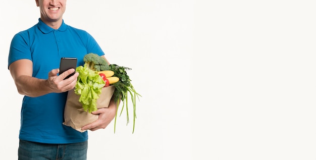 Smiley delivery man looking at smartphone while holding grocery bag