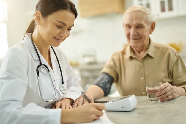 Smiley female doctor making notes while examining senior man sitting beside her