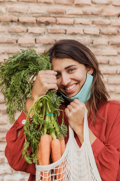 Smiley woman with face mask outdoors holding carrots