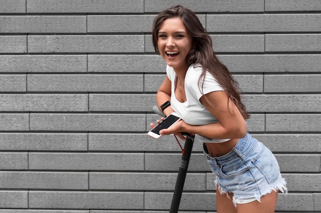 Photo smiley woman with phone in hand posing on electric scooter