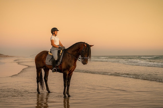 Un sorridente cavaliere di 21 anni in piedi sulla riva con il suo cavallo di fronte al mare