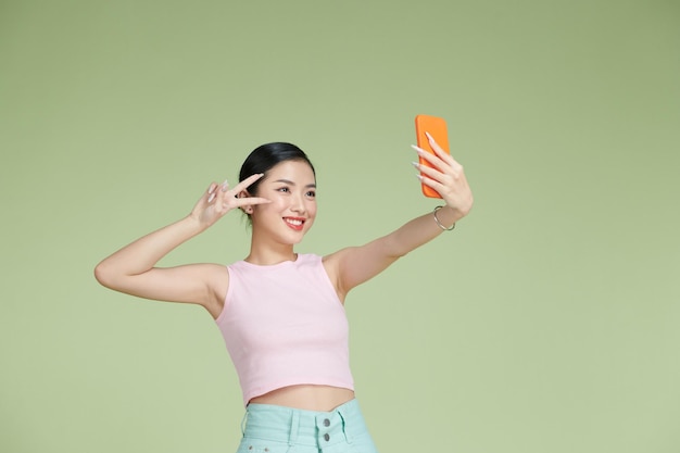 Smiling adorable asian female making selfie photo on smartphone with positive expression over green background