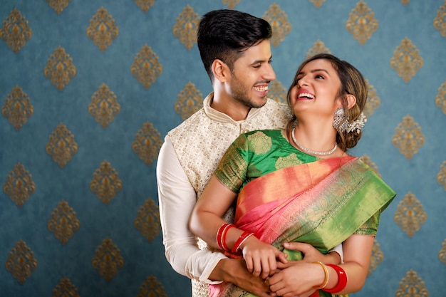 Улыбающаяся очаровательная пара, одетая в традиционную индийскую одежду, обнимается друг с другом перед камерой.
