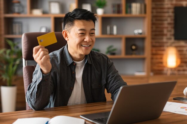 Smiling adult asian man show credit card work at computer use finance banking for online shopping