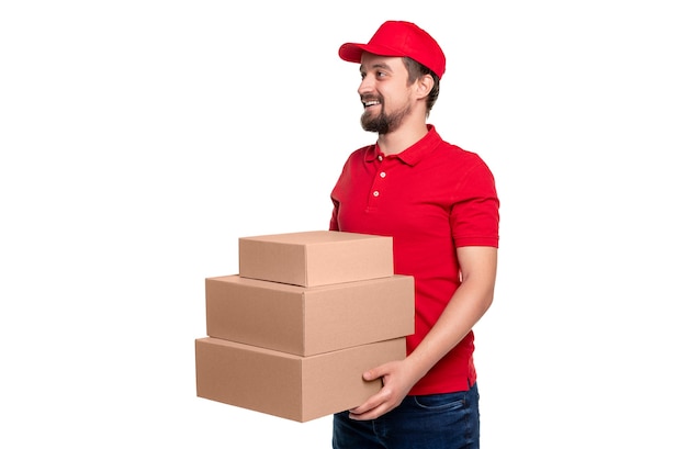 Smiling adult bearded male courier in red shirt and cap delivering stack of parcels in carton packages