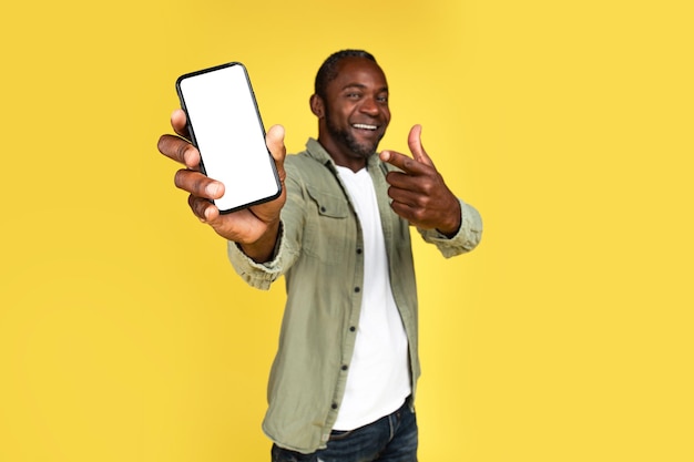 Smiling adult black guy in casual rejoices to online victory show finger at smartphone with blank screen