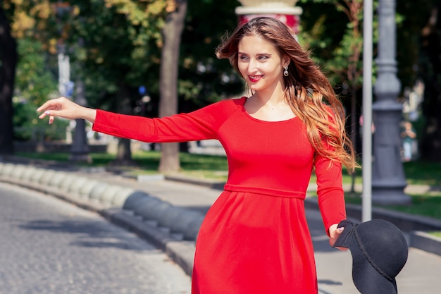 Smiling adult girl in red dress is catching taxi on the city street.