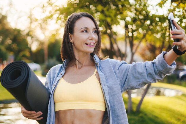 Smiling adult woman in sportswear standing in a grassy park taking a selfie on phone after doing yoga Fit woman jogging outdoor Happy lifestyle Healthy lifestyle sport