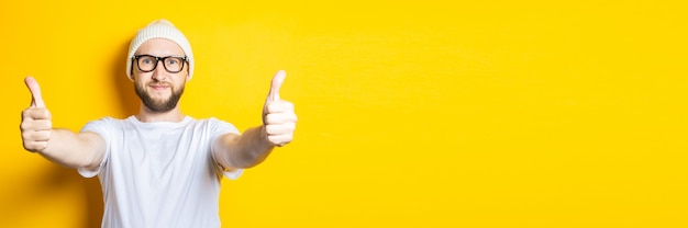 Smiling affable young guy with a beard in a cap and glasses shows a gesture with his hands