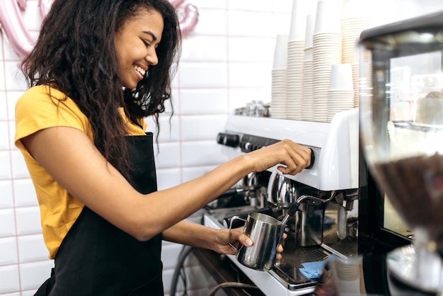 ユニフォームを着た笑顔のアフリカ系アメリカ人女性バリスタがコーヒーショップのビジターのためにコーヒーを作りますカフェの小規模ビジネスコンセプト