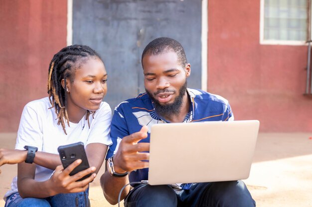 Smiling African American friends meet together at school holding phone and laptop device use modern technologies for entertainment