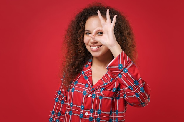 Smiling african american girl in pajamas homewear rest at home isolated on red background. relax good mood lifestyle concept. showing ok gesture hold hand near eye, imitating eyeglasses or binoculars.