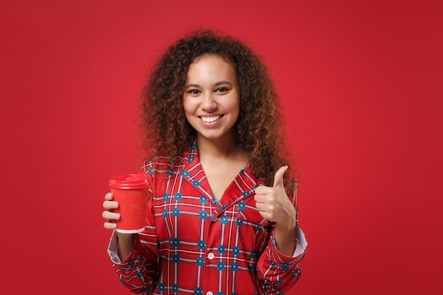 Smiling african american girl in pajamas homewear resting at\
home isolated on red background. relax good mood lifestyle concept.\
mock up copy space. hold paper cup of coffee or tea showing thumb\
up.