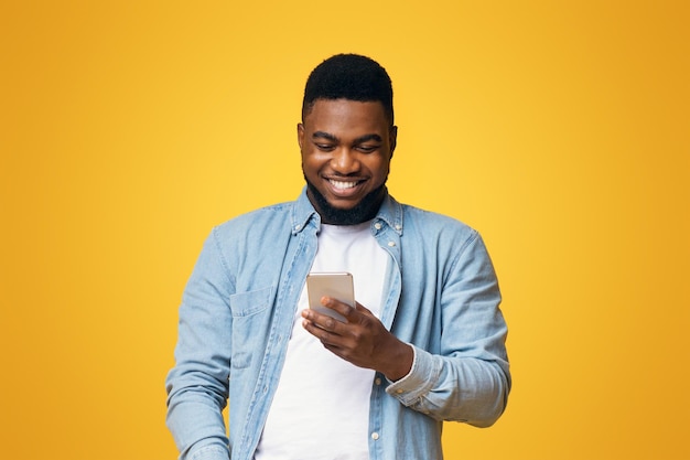 コピー付きの黄色いスタジオの背景でスマートフォンでメッセージを読んでいる笑顔のアフリカ系アメリカ人男性