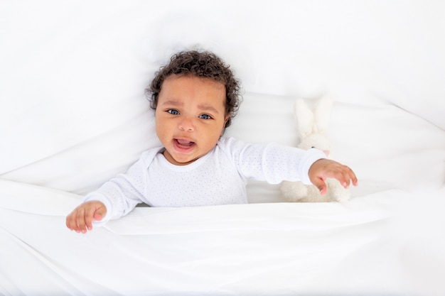 Smiling African-American little baby under a blanket with a toy on a white bed for sleeping.