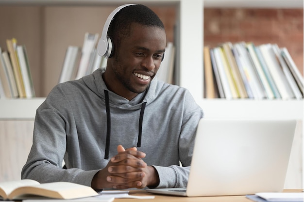 Smiling african American male sit at office desk wearing headphones watching online training course excited black guy in earphones enjoy learning webinar or tutorial on laptop at home