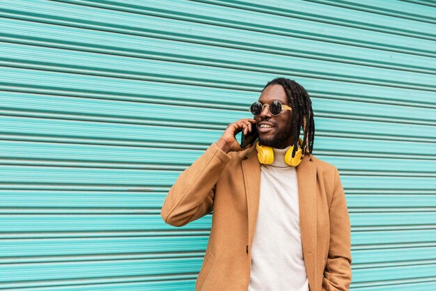 Smiling african american man in dreadlocks with stylish sunglasses with headphones around his neck on blue background talking by mobile phone