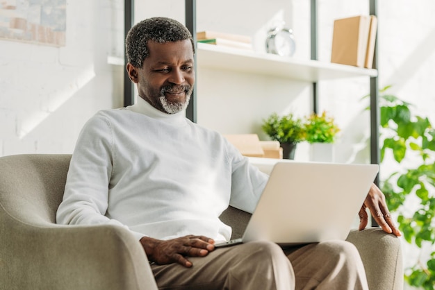 Smiling african american man sitting in armchair and using laptop