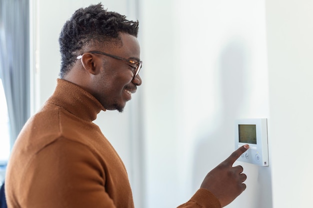 Улыбающийся афроамериканец, использующий современный контроллер системы умного дома на стене, положительный молодой человек переключает температуру на термостате или активирует охранную сигнализацию в квартире