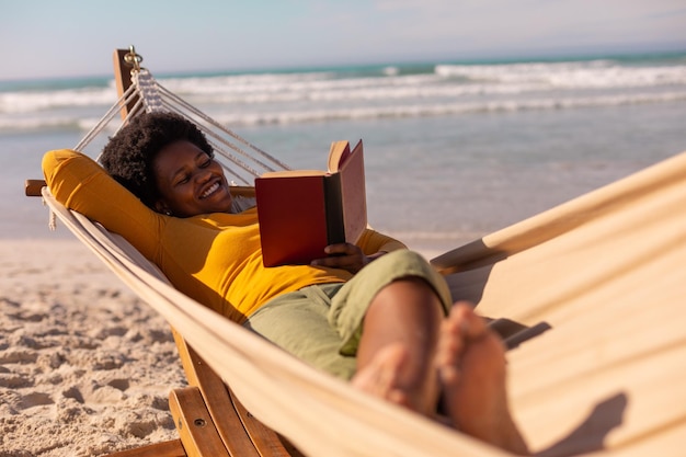 Foto donna matura afroamericana sorridente che legge un libro mentre giace su un'amaca contro il mare in estate