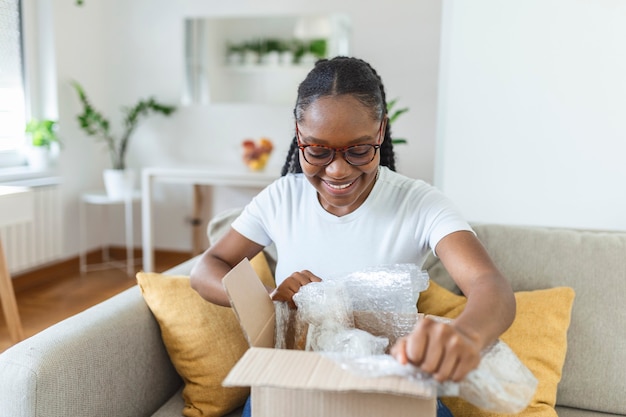 Smiling african American millennial girl sit on sofa at home open cardboard delivery package shopping online, excited black young woman unpack unwrap parcel buying good via internet or web