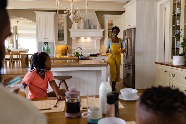 smiling african american mother with breakfast walking towards daughter at dining table. unaltered, lifestyle, family, love, togetherness, childhood, food, morning and home concept.