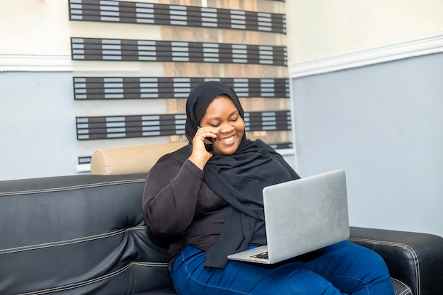 Sorridente afroamericano plus size donna musulmana che parla sul suo telefono cellulare facendo una chiamata d'affari seduto sul divano in soggiorno con computer portatile femmina che lavora in remoto