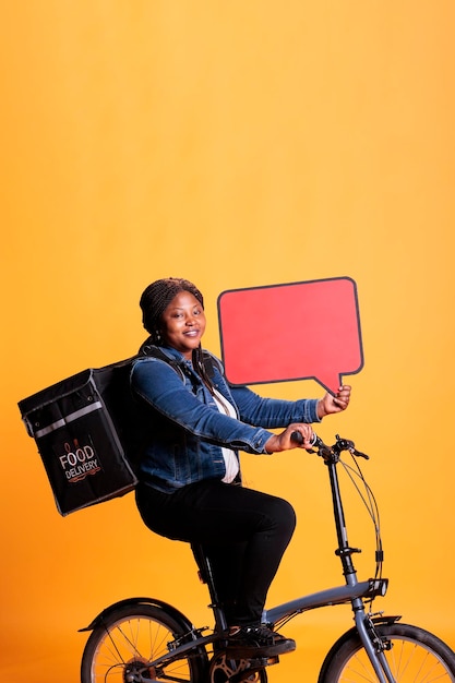 Smiling african american restaurant employee holding red speech bubble promoting takeaway food service while delivering fast food order to client using bike as transportation. Takeout concept