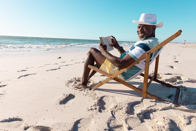 Sorridente uomo anziano afroamericano che indossa un cappello che legge un libro sulla sedia a sdraio in spiaggia sotto il cielo limpido. copia spazio, hobby, inalterato, stile di vita, vacanze, pensione, relax e concetto di natura.