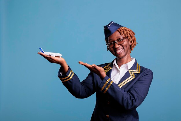 Smiling african american stewardess pointing at commercial\
airplane holding in hand for airline advertising. flight attendant\
wearing crew members uniform in studio shot.