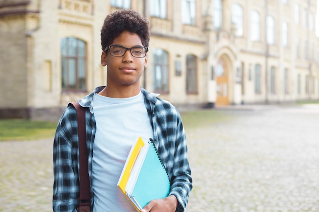 Smiling African American student with glasses and with books near college. Portrait of a happy black young man standing on a university. 