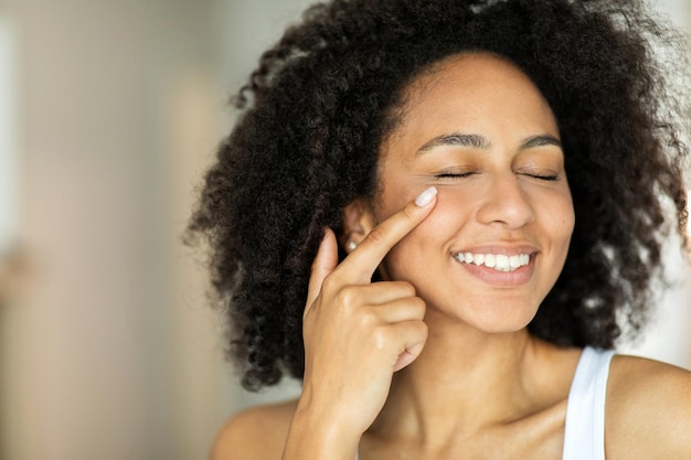 Donna afroamericana sorridente che applica crema idratante per il viso con gli occhi chiusi