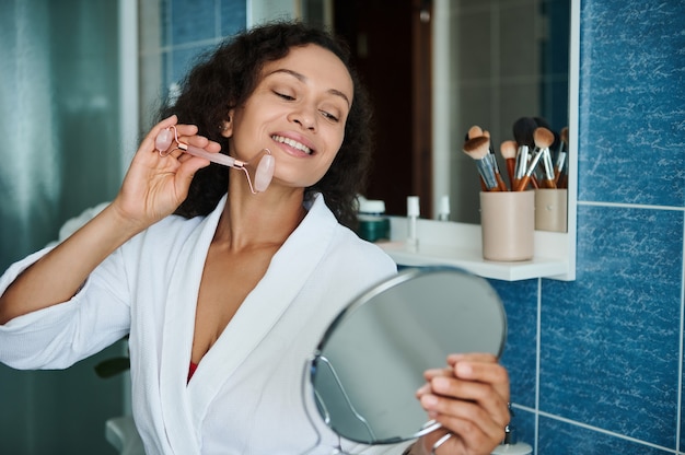 Smiling african american woman massaging her face with a jade roller massager and looking at her reflection in a small round cosmetic mirror