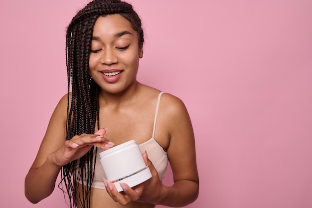 Smiling African American woman in underclothes posing with a jar with beauty cosmetic product and applying it, posing against a pink colored background