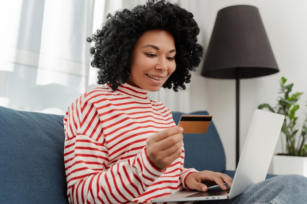 Smiling African American woman using laptop holding credit card shopping online ordering food