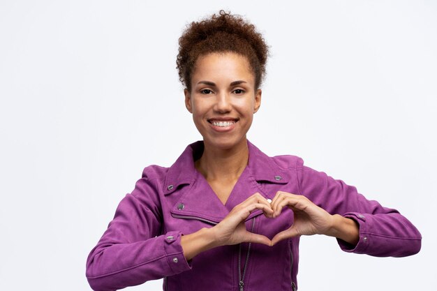 Smiling African American woman wearing purple jacket showing heart shape isolated on background