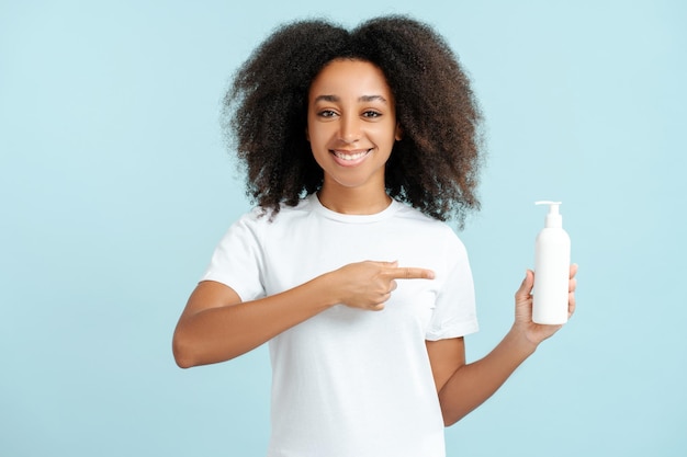 smiling African American woman with curly hair in white t shirt holding shampoo bottle mockup