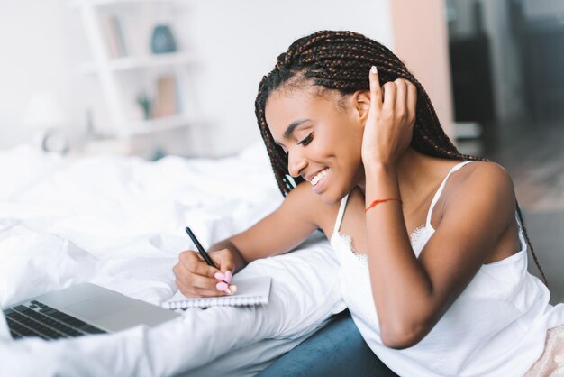 Smiling african american woman with laptop in bed writing in notepad